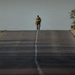 A single figure in military/tactical gear walks away from the camera along the center line of a straight, empty road. The person is carrying a large backpack with a prominent yellow-green reflective strip. The image uses shallow depth of field, with the foreground road blurred and the subject in sharp focus near the horizon.