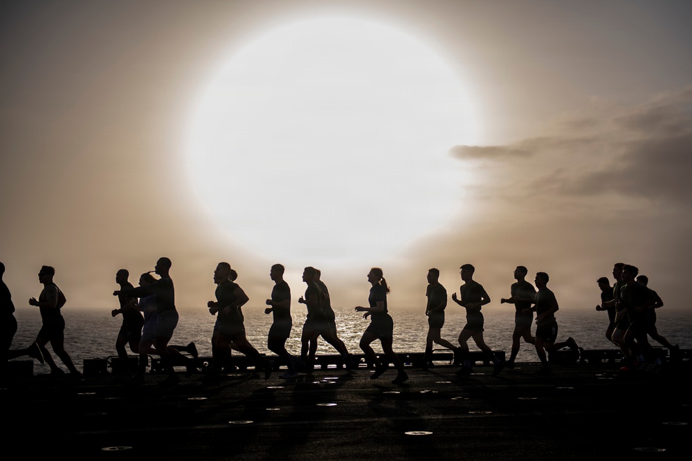 A group of people are running in silhouette against a large, glowing sun partially obscured by haze or fog, with an ocean horizon visible in the background. The scene appears to take place on a flat, dark surface — likely the flight deck of a naval vessel. The dramatic backlighting creates a striking contrast between the dark figures and the luminous sky.