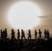 A group of people are running in silhouette against a large, glowing sun partially obscured by haze or fog, with an ocean horizon visible in the background. The scene appears to take place on a flat, dark surface — likely the flight deck of a naval vessel. The dramatic backlighting creates a striking contrast between the dark figures and the luminous sky.