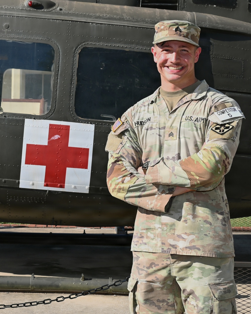 A U.S. Army Sergeant named Hudson stands with arms crossed in front of a military medical helicopter marked with a Red Cross symbol. He is wearing Operational Camouflage Pattern (OCP) uniform with visible rank insignia, name tape, U.S. Army tape, and a 'Competitor 5' tab on his right sleeve. The soldier is smiling and appears to be posing for an official or promotional photograph.