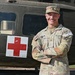 A U.S. Army Sergeant named Hudson stands with arms crossed in front of a military medical helicopter marked with a Red Cross symbol. He is wearing Operational Camouflage Pattern (OCP) uniform with visible rank insignia, name tape, U.S. Army tape, and a 'Competitor 5' tab on his right sleeve. The soldier is smiling and appears to be posing for an official or promotional photograph.
