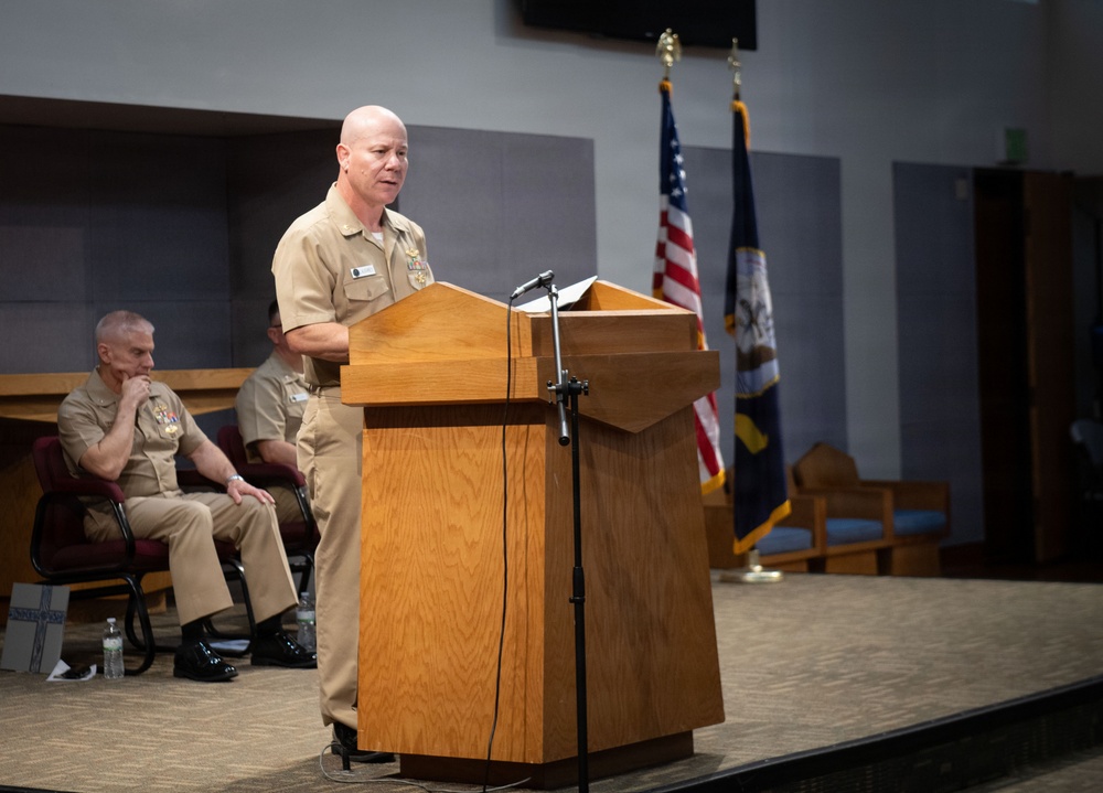 A U.S. Navy officer in Service Khaki uniform stands at a wooden podium delivering a speech at a formal military ceremony. Two other Navy personnel are seated on stage behind the speaker. The American flag and a Navy command flag are displayed in the background.