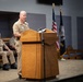 A U.S. Navy officer in Service Khaki uniform stands at a wooden podium delivering a speech at a formal military ceremony. Two other Navy personnel are seated on stage behind the speaker. The American flag and a Navy command flag are displayed in the background.