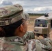 A U.S. Army soldier named Alvarado, wearing Operational Camouflage Pattern (OCP) uniform with a U.S. flag patch, is operating a tan-colored military electronic device in an open field setting. The soldier is looking through or aiming the device, which appears to be a targeting or signal detection system mounted on a tripod. The setting appears to be a training range or field exercise environment.