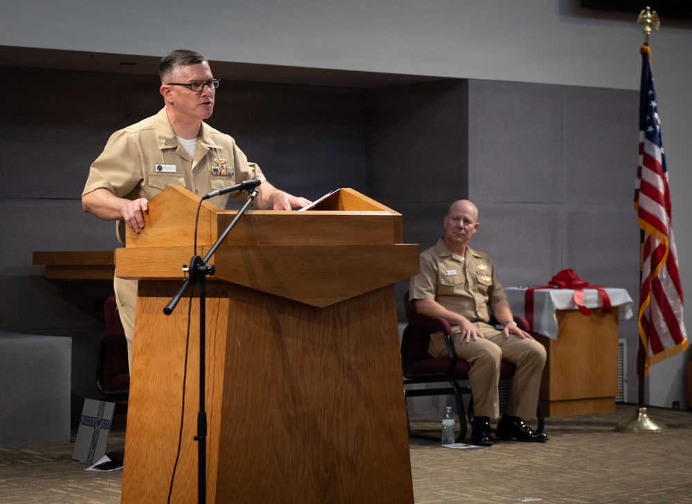 A U.S. Navy officer in tan service uniform is delivering a speech at a wooden podium equipped with a microphone. A second Navy officer is seated in the background, and an American flag is displayed to the right. The setting appears to be an official military ceremony or formal event held indoors.