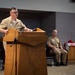 A U.S. Navy officer in tan service uniform is delivering a speech at a wooden podium equipped with a microphone. A second Navy officer is seated in the background, and an American flag is displayed to the right. The setting appears to be an official military ceremony or formal event held indoors.