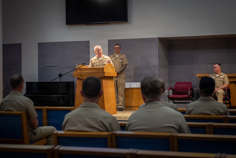 A military officer in tan/khaki service uniform is speaking at a wooden podium in what appears to be a chapel or auditorium setting. Several uniformed personnel are seated in pews facing the speaker, while two additional uniformed individuals stand and sit on the stage behind the podium. The setting and uniforms are consistent with a U.S. Navy formal ceremony or briefing.