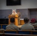 A military officer in tan/khaki service uniform is speaking at a wooden podium in what appears to be a chapel or auditorium setting. Several uniformed personnel are seated in pews facing the speaker, while two additional uniformed individuals stand and sit on the stage behind the podium. The setting and uniforms are consistent with a U.S. Navy formal ceremony or briefing.