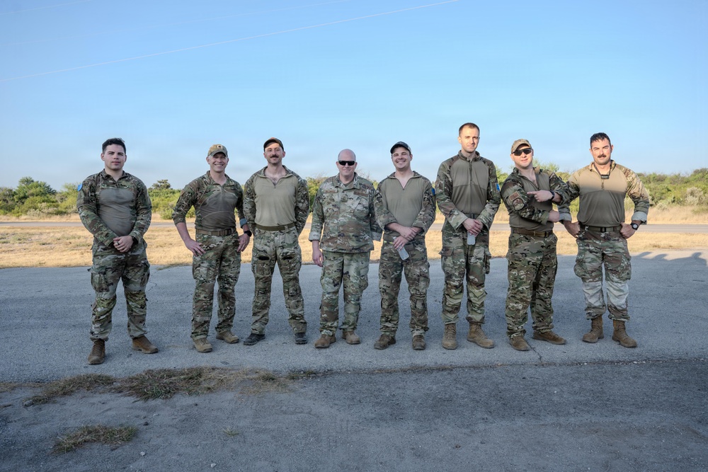 A group of nine military personnel in camouflage uniforms pose together for a group photograph outdoors. They are standing on a paved surface with dry grassland and sparse vegetation visible in the background under a clear blue sky. The individuals appear to be from a mixed unit or multinational team based on slight variations in their uniform patterns and colors.