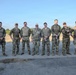 A group of nine military personnel in camouflage uniforms pose together for a group photograph outdoors. They are standing on a paved surface with dry grassland and sparse vegetation visible in the background under a clear blue sky. The individuals appear to be from a mixed unit or multinational team based on slight variations in their uniform patterns and colors.