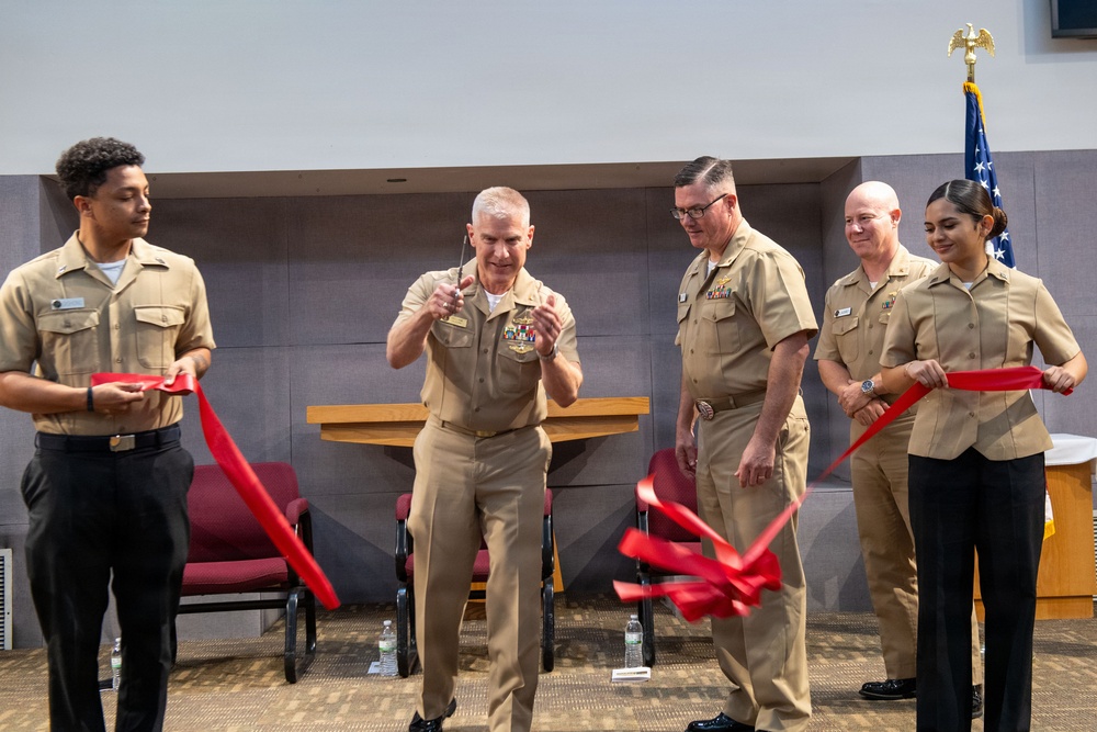 Five U.S. Navy personnel in tan service uniforms are participating in a ribbon-cutting ceremony in what appears to be an indoor military facility. A senior officer in the center, wearing multiple service ribbons and medals, has just cut a red ribbon while flanked by colleagues holding the ribbon ends. An American flag with an eagle finial is visible in the background.