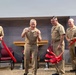Five U.S. Navy personnel in tan service uniforms are participating in a ribbon-cutting ceremony in what appears to be an indoor military facility. A senior officer in the center, wearing multiple service ribbons and medals, has just cut a red ribbon while flanked by colleagues holding the ribbon ends. An American flag with an eagle finial is visible in the background.