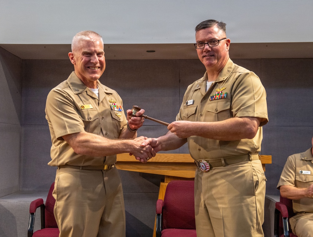 Two U.S. Navy officers in khaki service uniforms are shaking hands while jointly holding a ceremonial gavel, indicating a formal change of command or leadership transition ceremony. Both officers display multiple service ribbons and warfare qualification badges on their uniforms. The setting appears to be an indoor auditorium or conference room with red chairs visible in the background.