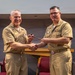 Two U.S. Navy officers in khaki service uniforms are shaking hands while jointly holding a ceremonial gavel, indicating a formal change of command or leadership transition ceremony. Both officers display multiple service ribbons and warfare qualification badges on their uniforms. The setting appears to be an indoor auditorium or conference room with red chairs visible in the background.