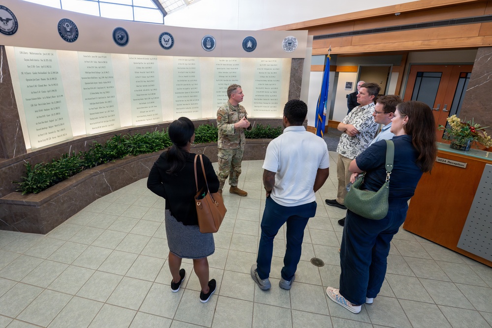 A military service member in camouflage uniform appears to be briefing or giving a tour to a small group of civilians in what appears to be a military installation lobby or memorial hall. The wall behind them displays multiple panels with names and dates, likely a memorial or honor roll, adorned with military branch seals including the Navy, Air Force, Space Force, and Coast Guard. A blue military flag is visible in the background near a reception desk labeled 'OO2A Reception'.