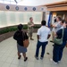 A military service member in camouflage uniform appears to be briefing or giving a tour to a small group of civilians in what appears to be a military installation lobby or memorial hall. The wall behind them displays multiple panels with names and dates, likely a memorial or honor roll, adorned with military branch seals including the Navy, Air Force, Space Force, and Coast Guard. A blue military flag is visible in the background near a reception desk labeled 'OO2A Reception'.