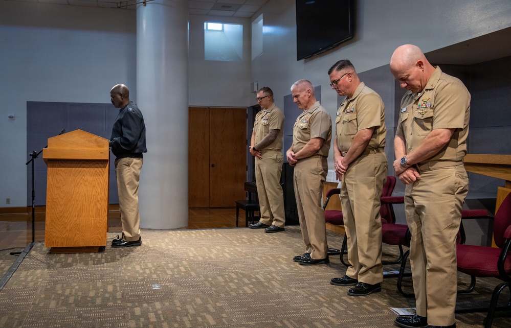 A formal military ceremony or prayer service is taking place in what appears to be a chapel or meeting room. A civilian or chaplain in dark clothing stands at a wooden podium while four U.S. Navy personnel in tan Service Khaki uniforms stand with bowed heads in a posture of prayer or reverence. The officers display ribbons and insignia consistent with senior Navy ranks.