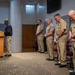 A formal military ceremony or prayer service is taking place in what appears to be a chapel or meeting room. A civilian or chaplain in dark clothing stands at a wooden podium while four U.S. Navy personnel in tan Service Khaki uniforms stand with bowed heads in a posture of prayer or reverence. The officers display ribbons and insignia consistent with senior Navy ranks.