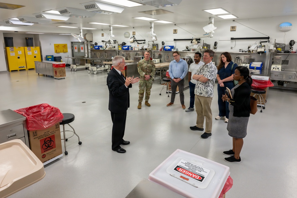 A group of approximately seven people are gathered in what appears to be a medical examiner's office or forensic laboratory facility. A man in a dark suit appears to be briefing or presenting to the group, which includes a military personnel in camouflage uniform and several civilians. The room contains medical/forensic equipment, biohazard waste containers, and numbered workstations along the walls.