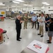 A group of approximately seven people are gathered in what appears to be a medical examiner's office or forensic laboratory facility. A man in a dark suit appears to be briefing or presenting to the group, which includes a military personnel in camouflage uniform and several civilians. The room contains medical/forensic equipment, biohazard waste containers, and numbered workstations along the walls.