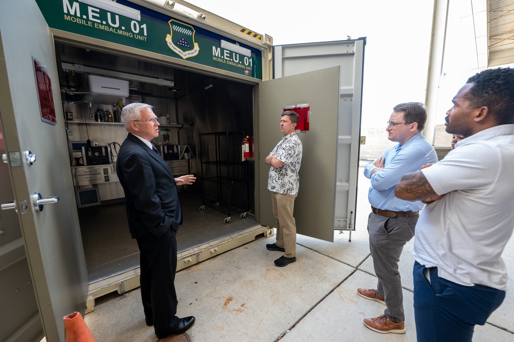 Four men are gathered outside an open shipping container labeled 'M.E.U. 01 - Mobile Embalming Unit,' which appears to be a military mortuary affairs asset. An older man in a dark suit appears to be briefing or presenting the unit to three other men in civilian attire. The container interior is visible and contains shelving, equipment, and supplies.