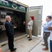 Four men are gathered outside an open shipping container labeled 'M.E.U. 01 - Mobile Embalming Unit,' which appears to be a military mortuary affairs asset. An older man in a dark suit appears to be briefing or presenting the unit to three other men in civilian attire. The container interior is visible and contains shelving, equipment, and supplies.
