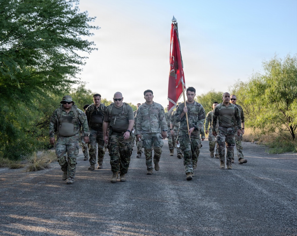 A group of military personnel in camouflage uniforms are conducting a ruck march or formation walk along a paved road in what appears to be a desert or arid environment. One soldier carries a red unit guidon flag with a spear-tipped staff. The group is moving toward the camera in a loose formation during daylight hours.