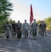 A group of military personnel in camouflage uniforms are conducting a ruck march or formation walk along a paved road in what appears to be a desert or arid environment. One soldier carries a red unit guidon flag with a spear-tipped staff. The group is moving toward the camera in a loose formation during daylight hours.