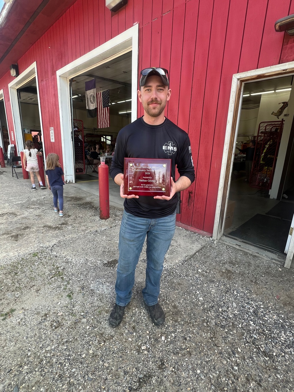 A man wearing a black EMS long-sleeve shirt and a baseball cap stands in front of a red building, holding a wooden award plaque. The plaque appears to be a 'Denali Borough Mayor's Community Service Award' presented to 'Nathan Grimes' for 2026. The setting appears to be a fire station or emergency services facility, with an American flag and equipment visible in the background.