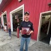 A man wearing a black EMS long-sleeve shirt and a baseball cap stands in front of a red building, holding a wooden award plaque. The plaque appears to be a 'Denali Borough Mayor's Community Service Award' presented to 'Nathan Grimes' for 2026. The setting appears to be a fire station or emergency services facility, with an American flag and equipment visible in the background.