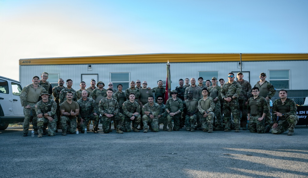 A large group of military personnel, approximately 35-40 individuals, pose for a formal unit photograph in front of an ATCO modular building. The group is arranged in two rows — standing in the back and kneeling in the front — wearing various camouflage uniforms. A military guidon (unit flag) is visible in the center background, and a partially visible sign on the right references an Engineer (EN) Battalion.