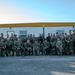 A large group of military personnel, approximately 35-40 individuals, pose for a formal unit photograph in front of an ATCO modular building. The group is arranged in two rows — standing in the back and kneeling in the front — wearing various camouflage uniforms. A military guidon (unit flag) is visible in the center background, and a partially visible sign on the right references an Engineer (EN) Battalion.