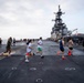 Military personnel are conducting a running event or physical fitness exercise on the flight deck of a large amphibious assault ship. The deck is marked with orange traffic cones to guide runners along a course, and at least one F-35B stealth aircraft is visible parked on the starboard side. The ship's island superstructure with radar and communication equipment is prominently visible in the background.