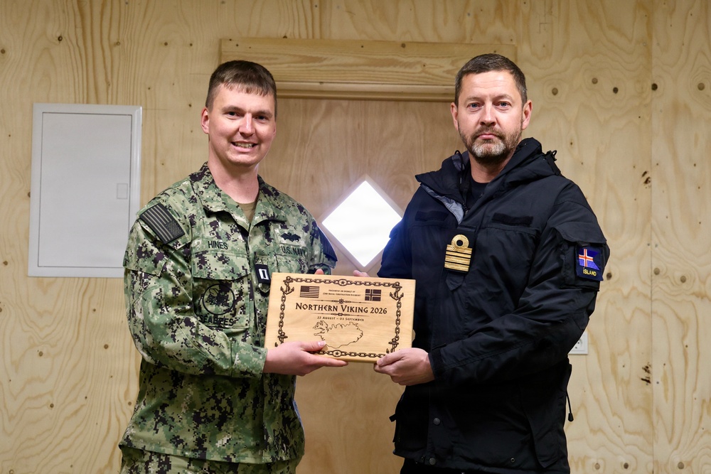Two military personnel are posing together while holding a wooden commemorative plaque for 'Northern Viking 2026,' a joint military exercise. The individual on the left is a U.S. Navy servicemember in digital camouflage (NWU Type III) with the name tape 'HINES' and a Seabee patch, while the individual on the right wears a black uniform with an 'ÍSLAND' (Iceland) patch and gold rank insignia. The ceremony appears to be a formal exchange or presentation between U.S. and Icelandic military representatives.