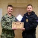 Two military personnel are posing together while holding a wooden commemorative plaque for 'Northern Viking 2026,' a joint military exercise. The individual on the left is a U.S. Navy servicemember in digital camouflage (NWU Type III) with the name tape 'HINES' and a Seabee patch, while the individual on the right wears a black uniform with an 'ÍSLAND' (Iceland) patch and gold rank insignia. The ceremony appears to be a formal exchange or presentation between U.S. and Icelandic military representatives.
