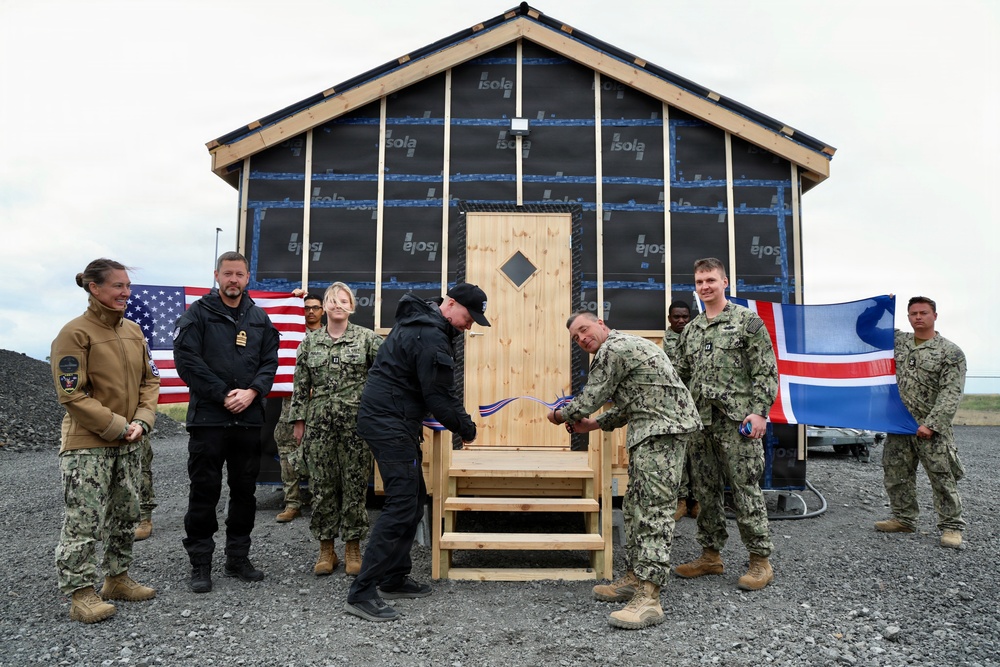 A ribbon-cutting ceremony is taking place in front of a newly constructed wooden building with 'Isola' brand weather barrier wrap visible on its exterior. Military personnel in U.S. Navy camouflage uniforms are gathered alongside civilians, with both an American flag and an Icelandic flag being held in the background. The ceremony appears to mark the completion or inauguration of a newly built structure.