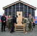 A ribbon-cutting ceremony is taking place in front of a newly constructed wooden building with 'Isola' brand weather barrier wrap visible on its exterior. Military personnel in U.S. Navy camouflage uniforms are gathered alongside civilians, with both an American flag and an Icelandic flag being held in the background. The ceremony appears to mark the completion or inauguration of a newly built structure.