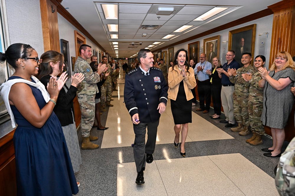A high-ranking military officer in a dark dress uniform with medals and insignia walks down a hallway corridor alongside a civilian woman, while a crowd of military personnel in camouflage uniforms and civilians applaud on both sides. The setting appears to be an official military building with portrait paintings lining the walls, suggesting a formal recognition or farewell ceremony. The atmosphere is celebratory, with smiling faces and enthusiastic applause from all attendees.