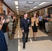 A high-ranking military officer in a dark dress uniform with medals and insignia walks down a hallway corridor alongside a civilian woman, while a crowd of military personnel in camouflage uniforms and civilians applaud on both sides. The setting appears to be an official military building with portrait paintings lining the walls, suggesting a formal recognition or farewell ceremony. The atmosphere is celebratory, with smiling faces and enthusiastic applause from all attendees.