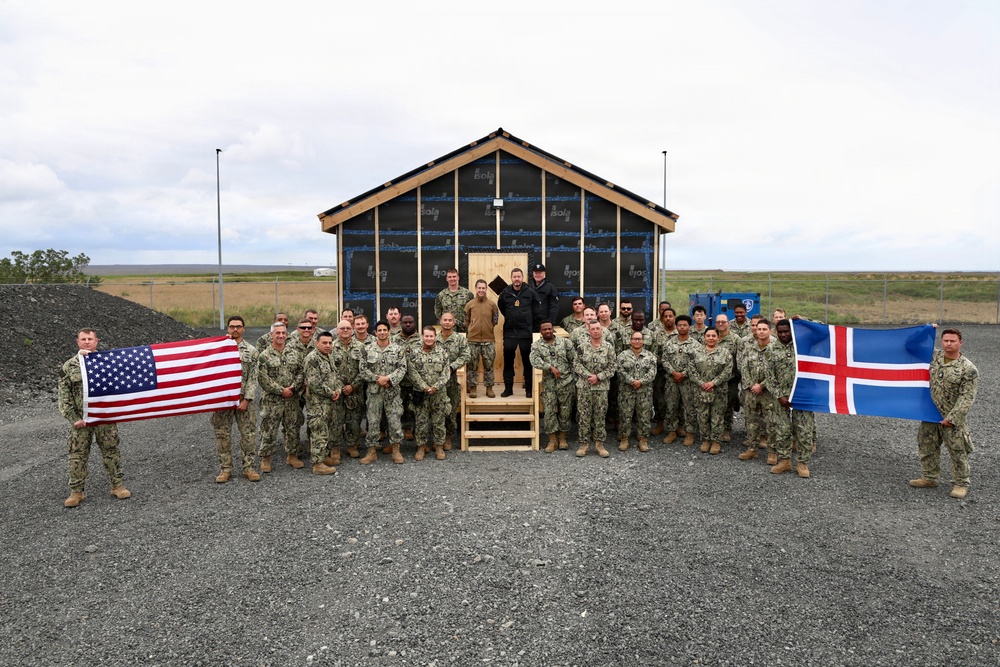 A large group of military personnel in camouflage uniforms pose for a group photograph in front of a newly constructed building frame covered with weather barrier wrap. Two flags are prominently displayed on either side of the group — the United States flag on the left and the Icelandic flag on the right. The setting appears to be a remote, flat terrain consistent with Iceland, suggesting a joint US-Icelandic military construction or engineering project.