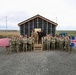 A large group of military personnel in camouflage uniforms pose for a group photograph in front of a newly constructed building frame covered with weather barrier wrap. Two flags are prominently displayed on either side of the group — the United States flag on the left and the Icelandic flag on the right. The setting appears to be a remote, flat terrain consistent with Iceland, suggesting a joint US-Icelandic military construction or engineering project.