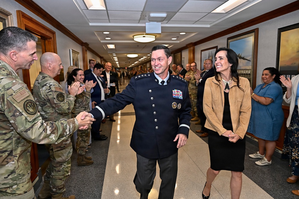 A senior military officer in a dark blue dress uniform with four-star insignia and numerous medals walks down a hallway flanked by applauding personnel and civilians, accompanied by a woman in civilian attire. The setting appears to be an official military building with framed aviation artwork on the walls. The scene is consistent with a formal military ceremony, such as a promotion, retirement, or change-of-command event.