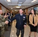 A senior military officer in a dark blue dress uniform with four-star insignia and numerous medals walks down a hallway flanked by applauding personnel and civilians, accompanied by a woman in civilian attire. The setting appears to be an official military building with framed aviation artwork on the walls. The scene is consistent with a formal military ceremony, such as a promotion, retirement, or change-of-command event.