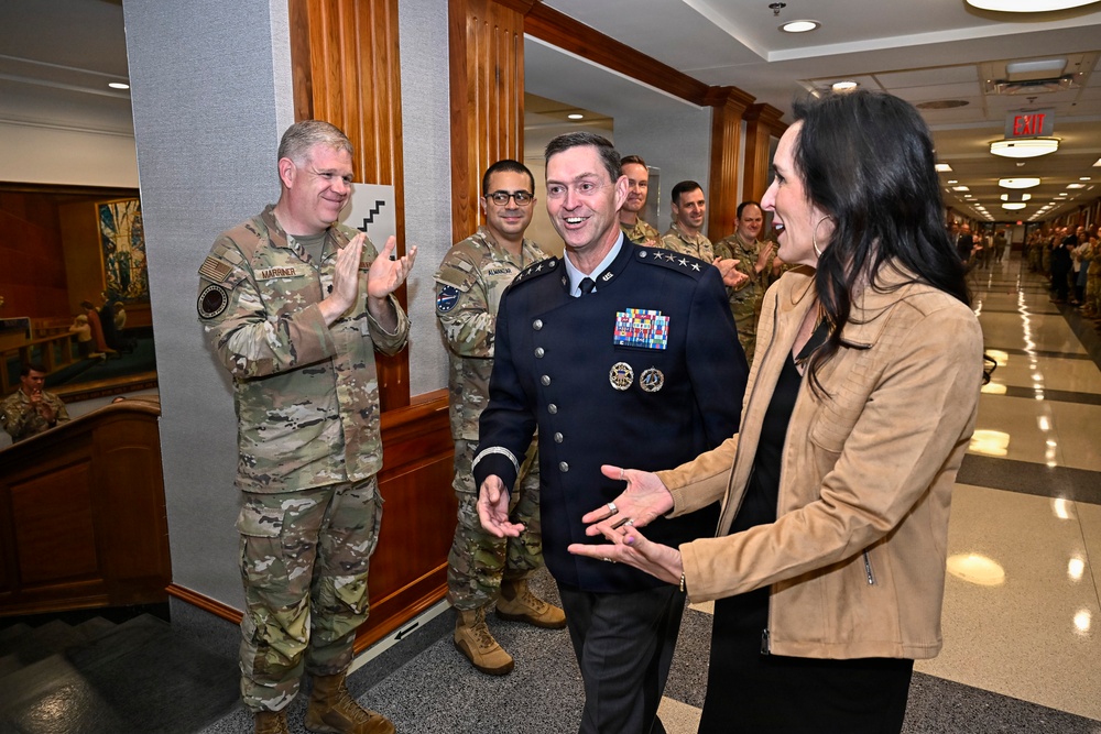 A formal military ceremony or reception is taking place in what appears to be a government or military building hallway. A senior U.S. Air Force officer in dress blues with multiple ribbons and two-star rank insignia is being greeted or congratulated by a civilian woman and surrounded by personnel in camouflage uniforms. The atmosphere appears celebratory, with personnel applauding in the background.