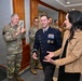 A formal military ceremony or reception is taking place in what appears to be a government or military building hallway. A senior U.S. Air Force officer in dress blues with multiple ribbons and two-star rank insignia is being greeted or congratulated by a civilian woman and surrounded by personnel in camouflage uniforms. The atmosphere appears celebratory, with personnel applauding in the background.