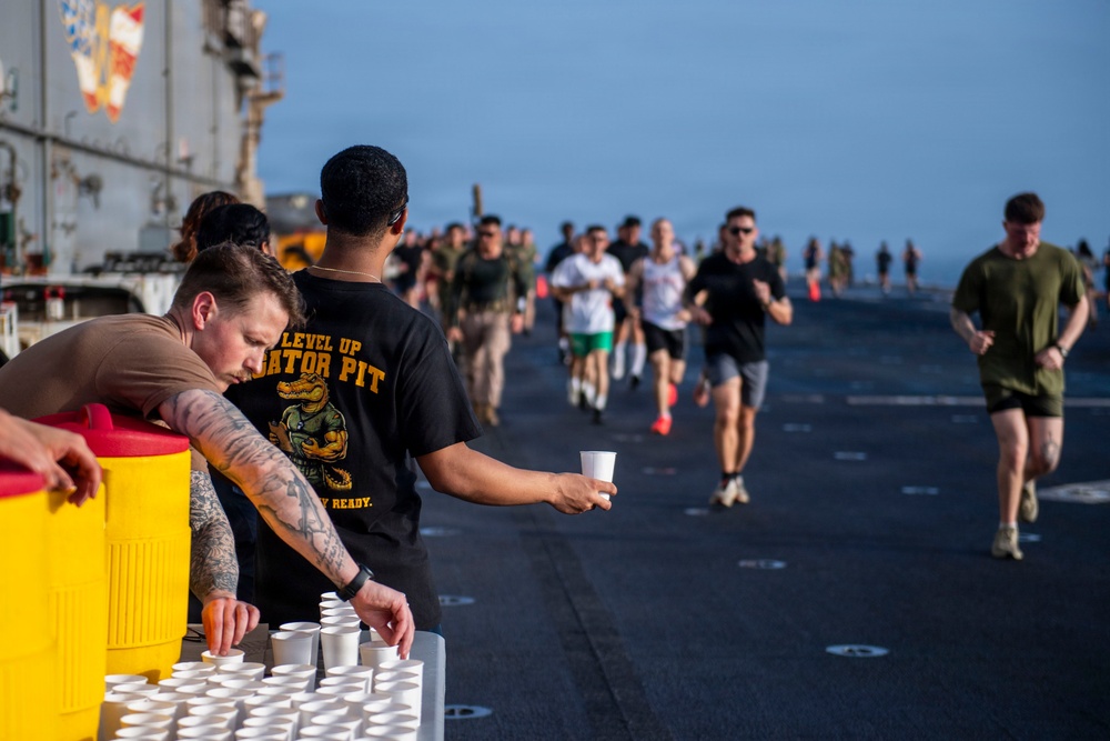 A group of military personnel are participating in a running event on what appears to be the flight deck of a naval vessel. Two volunteers man a water station in the foreground, handing out small paper cups to passing runners. The event resembles a 5K or fun run organized aboard a ship at sea.