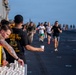 A group of military personnel are participating in a running event on what appears to be the flight deck of a naval vessel. Two volunteers man a water station in the foreground, handing out small paper cups to passing runners. The event resembles a 5K or fun run organized aboard a ship at sea.