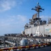 The image shows the flight deck of a U.S. Navy amphibious assault ship, identified by the number '4' painted on the island superstructure. Personnel are engaged in physical fitness activities, running along the flight deck while at sea. The ship displays safety markings including 'BEWARE OF JET BLAST AND ROTORS' and features a naval aviation insignia on the hull.