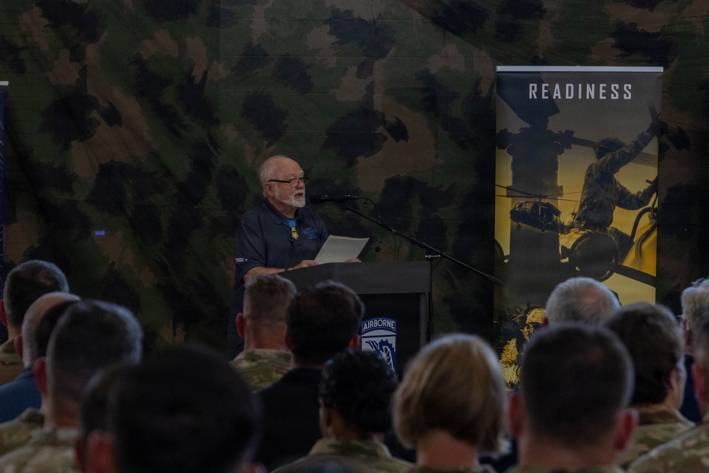 An older civilian man with white hair and a beard is speaking at a podium during a military event. The podium displays the 18th Airborne Corps insignia, and a 'READINESS' banner is visible in the background. An audience of military personnel in camouflage uniforms is seated facing the speaker.