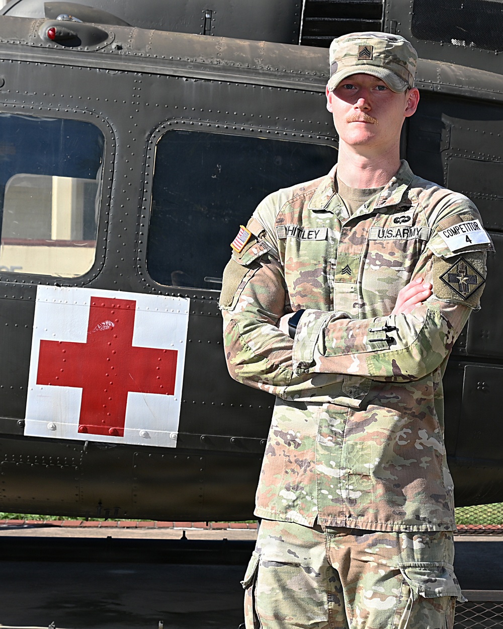 A U.S. Army Sergeant named Whitley stands with arms crossed in front of a military medical helicopter marked with a Red Cross symbol. He is wearing Operational Camouflage Pattern (OCP) uniform with visible rank insignia, name tape, U.S. Army tape, a unit patch, and a 'Competitor 4' armband. The image appears to be an official or promotional military photograph.